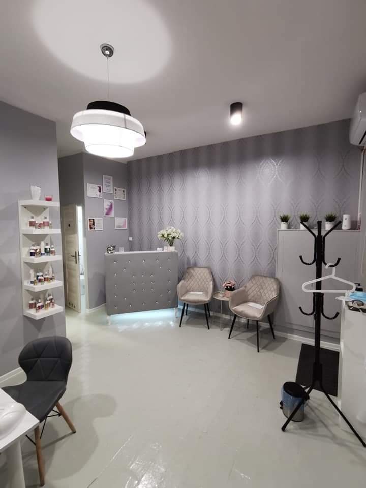 Jasne, szaro-białe wnętrze salonu kosmetycznego z recepcją obitą guzikami, dwoma fotelami, dekoracyjną tapetą i regałem z lakierami do paznokci.
