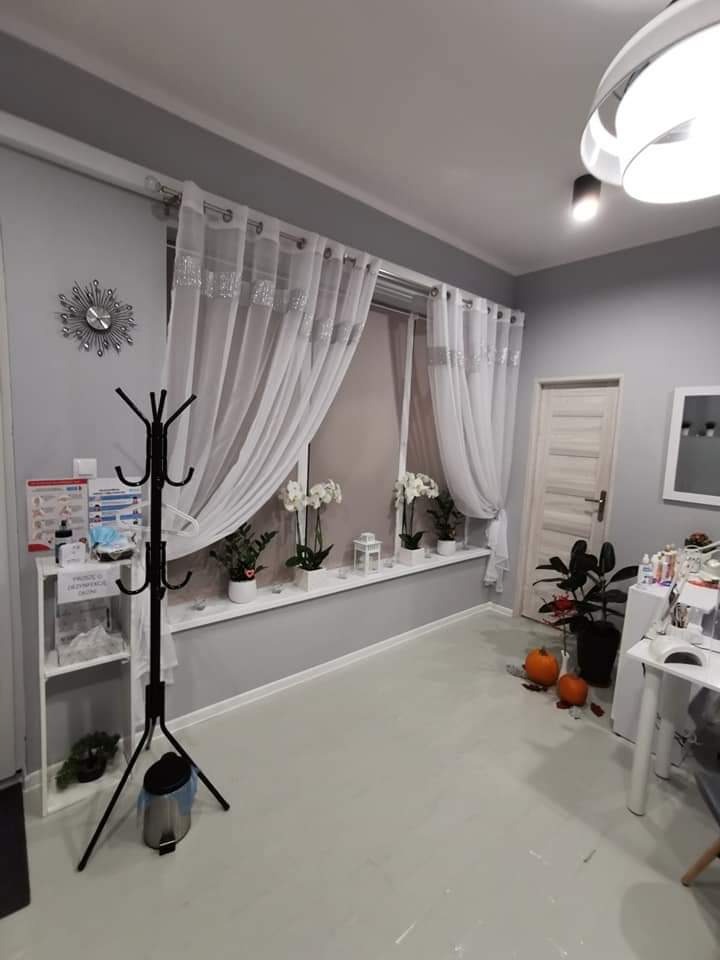 Jasne wnętrze salonu kosmetycznego z szarymi ścianami, białymi zasłonami i roślinami w oknach, uzupełnione dekoracjami sezonowymi w postaci dwóch małych dyń na podłodze.