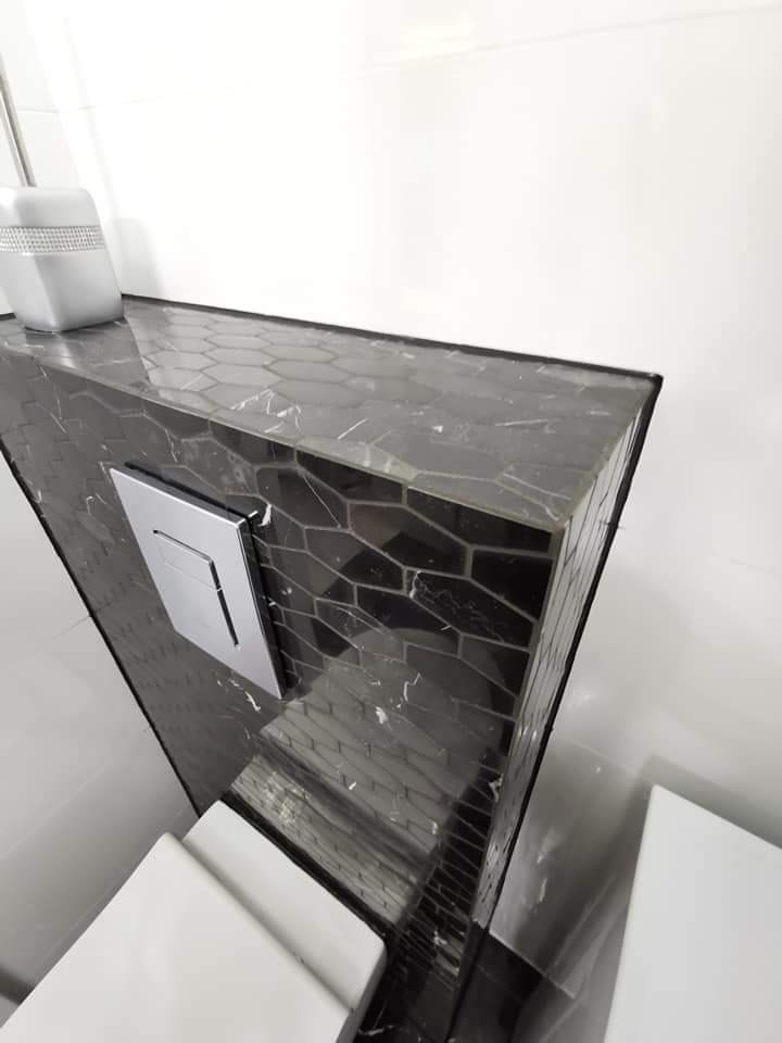 Fragment łazienki z zabudową stelaża WC wykończoną czarną mozaiką heksagonalną z białymi żyłkami, chromowany przycisk spłuczki, białe płytki ścienne i ceramiczny pojemnik na szczotkę do WC...