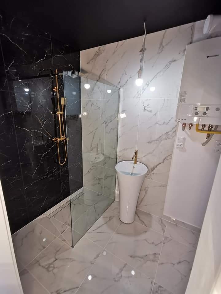 Nowoczesna łazienka z czarną ścianą prysznicową z marmurowym wzorem, szklaną kabiną prysznicową, wolnostojącą białą umywalką na postumencie z złotą armaturą, oraz biało-szarą marmurową podłogą...