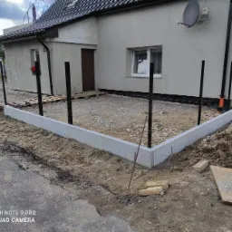 Przygotowanie podłoża pod taras: wylany betonowy fundament, widoczne czarne słupki, palety, piasek, ziemia i rozciągnięte sznurki wyznaczające granice. Budynek w tle.