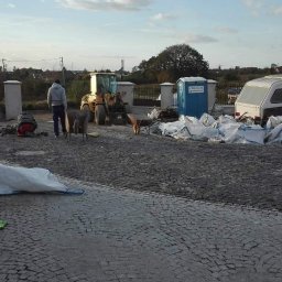 ggworkbruk - Układanie kostki granitowej na posesji z widocznym traktorem, toaletą przenośną i przyczepą kempingową w tle. Na pierwszym planie widoczne dwie owce.