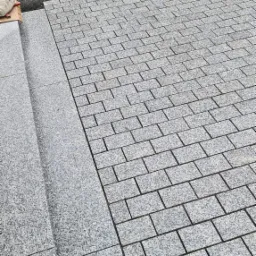 Ułożona z jasnoszarej kostki granitowej ścieżka przechodzi w dwustopniowe schody z płyt granitowych, w tle widoczny fragment przygotowywanego terenu pod dalsze prace brukarskie.