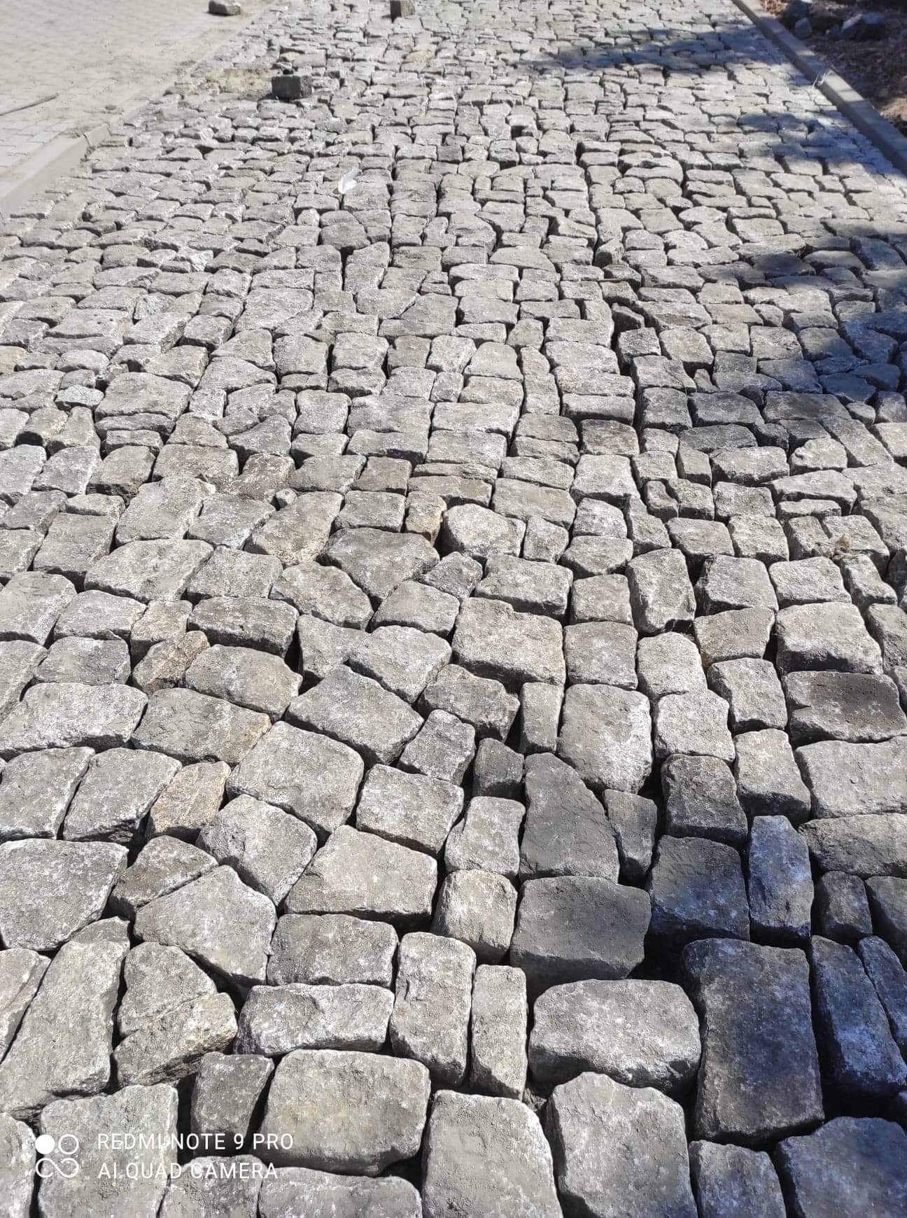 Szara nawierzchnia z nieregularnej kostki kamiennej ułożonej wzdłuż drogi, widok z góry z lekkim cieniem.