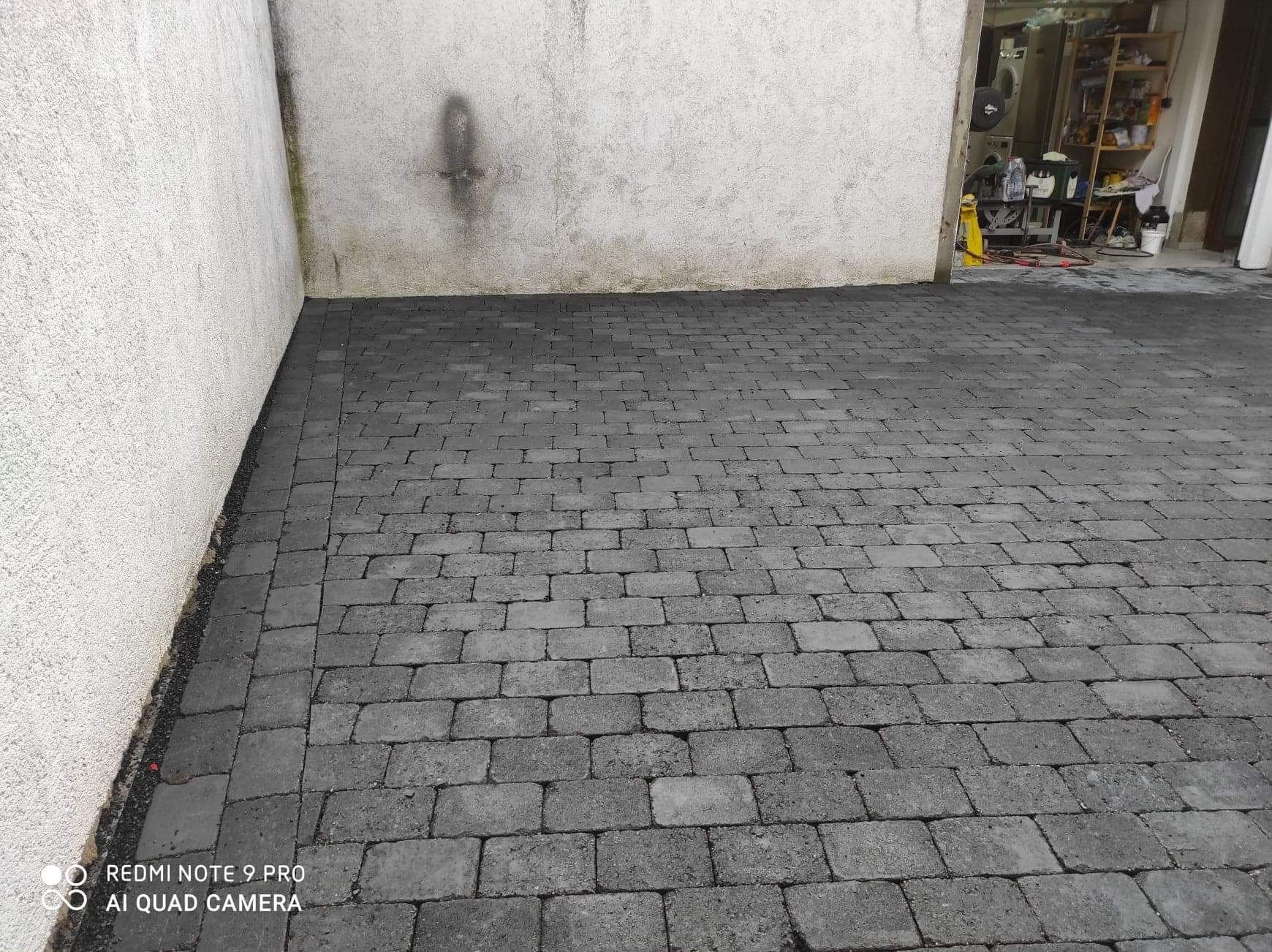 Ułożona szara kostka brukowa na podjeździe, widok z góry, ściana budynku po lewej stronie, widoczne pomieszczenie gospodarcze w tle po prawej.