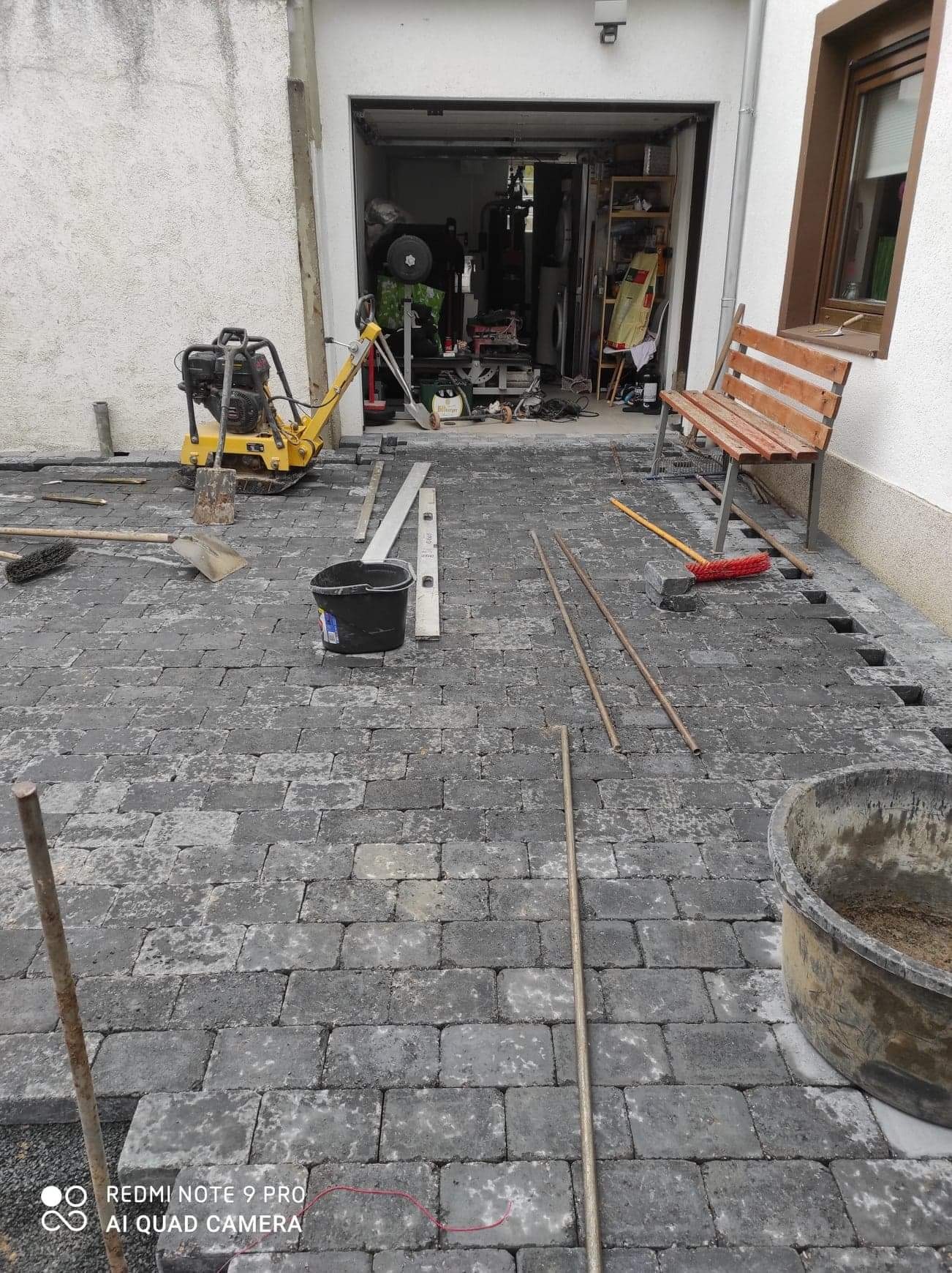 Układanie szarej kostki brukowej na posesji, widoczny ubijak wibracyjny, poziomica, rury, wiadro z zaprawą oraz częściowo ułożona nawierzchnia.