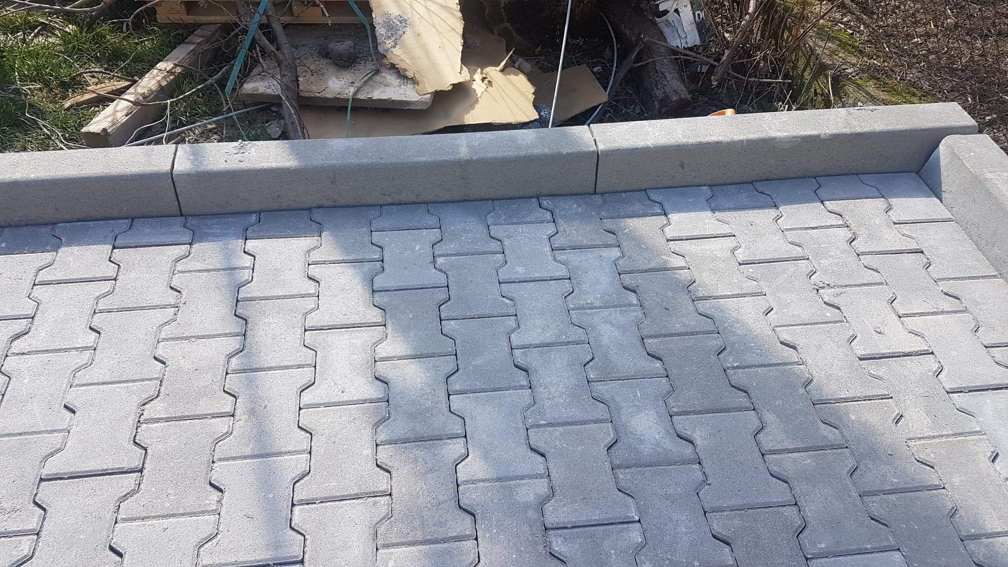 Ułożona szara kostka brukowa w kształcie litery H z obrzeżami, widoczne ślady piasku w spoinach, w tle gałęzie i resztki materiałów budowlanych.