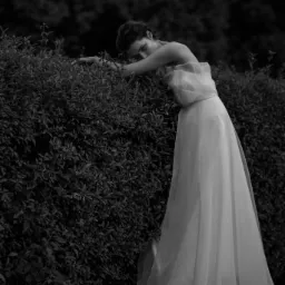 Kobieta w zwiewnej, jasnej sukni opiera się o gęsty, ciemny żywopłot w czarno-białej fotografii, z delikatnym spojrzeniem w stronę obiektywu.