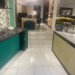 K&G MEBLE KINGA GNYLA - Nowoczesne wnętrze kuchni z wyspą o turkusowym wykończeniu, czarnym blatem i wbudowanym regałem na wino, widok na biuro w tle, białe marmurowe płytki podłogowe.