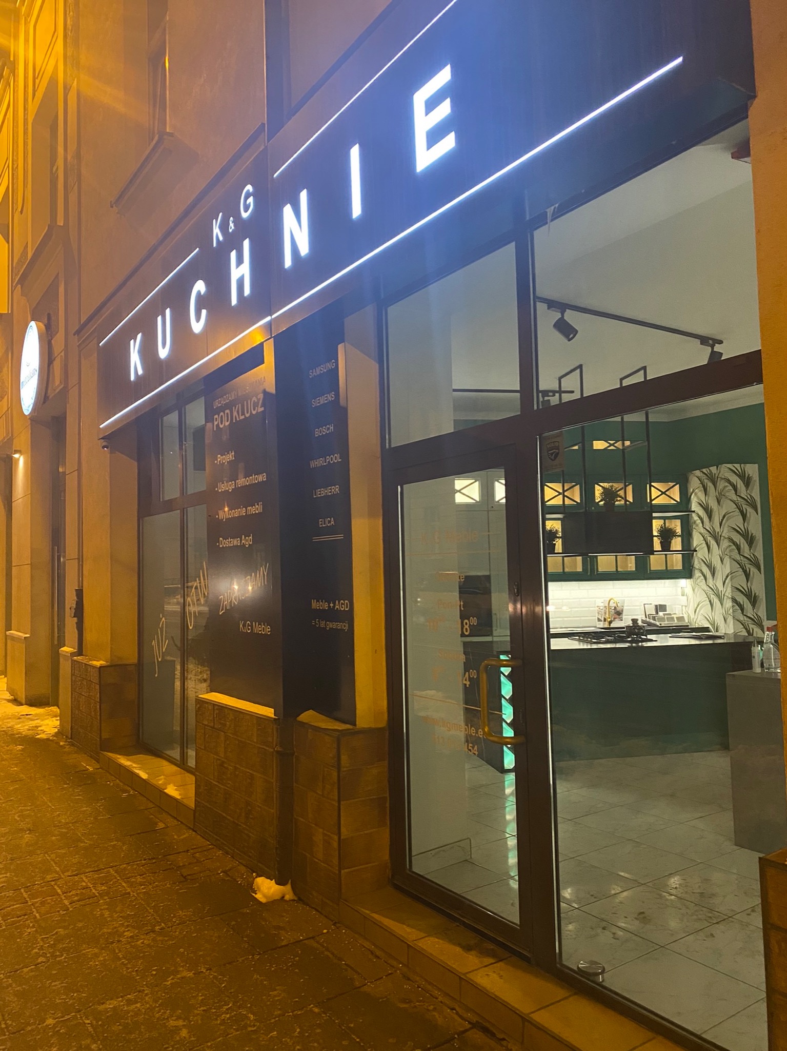 Fasada studia meblowego K&G MEBLE w Krakowie, z widocznym szyldem 'KUCHNIE' podświetlonym neonem, ofertą 'pod klucz' na szybie i ekspozycją wnętrza z zabudową kuchenną w odcieniach zieleni.