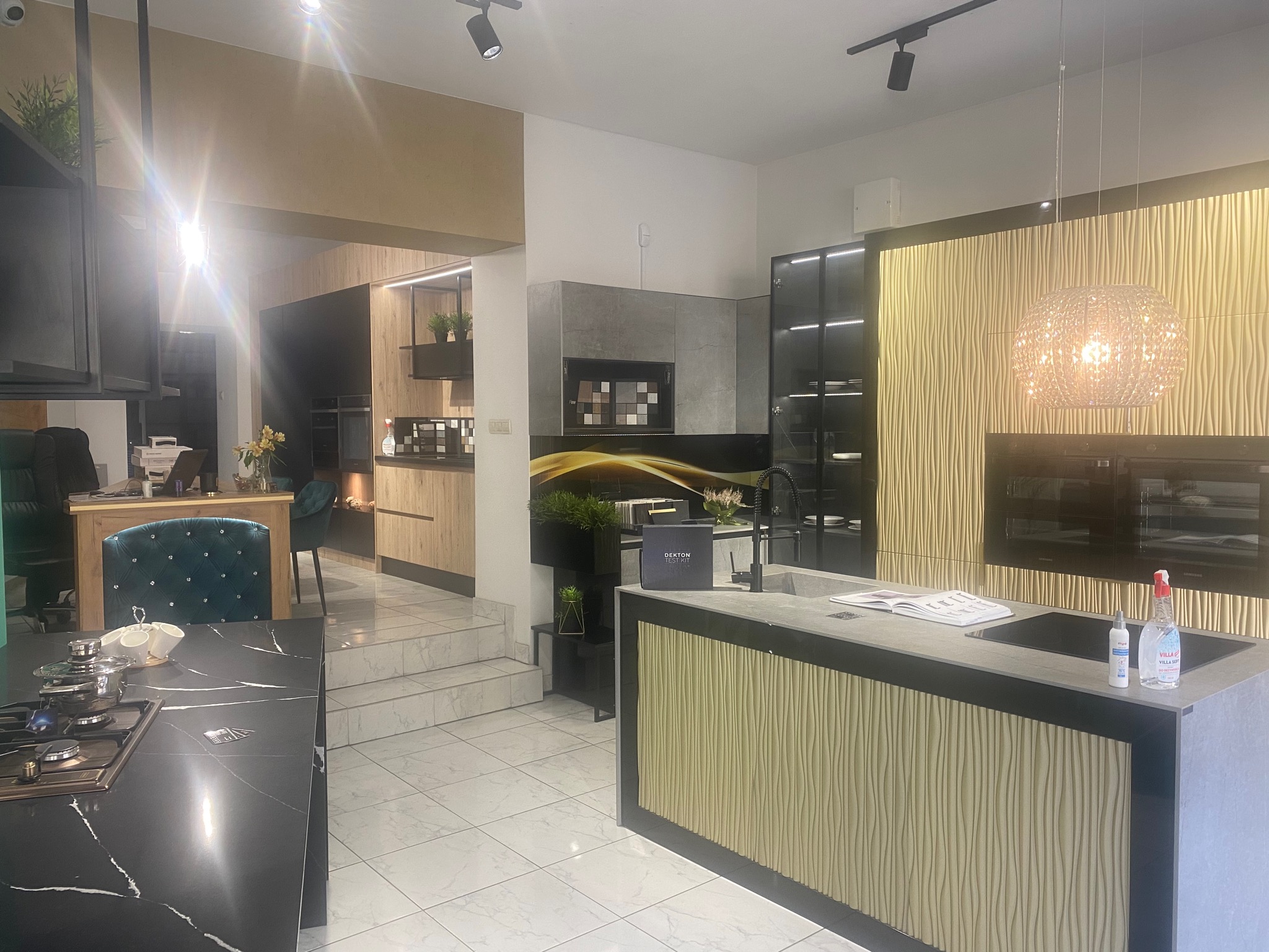 Salon ekspozycyjny z nowoczesną zabudową kuchenną w odcieniach drewna i szarości, z wyspą kuchenną z blatem z szarego spieku i dekoracyjnym oświetleniem wiszącym.