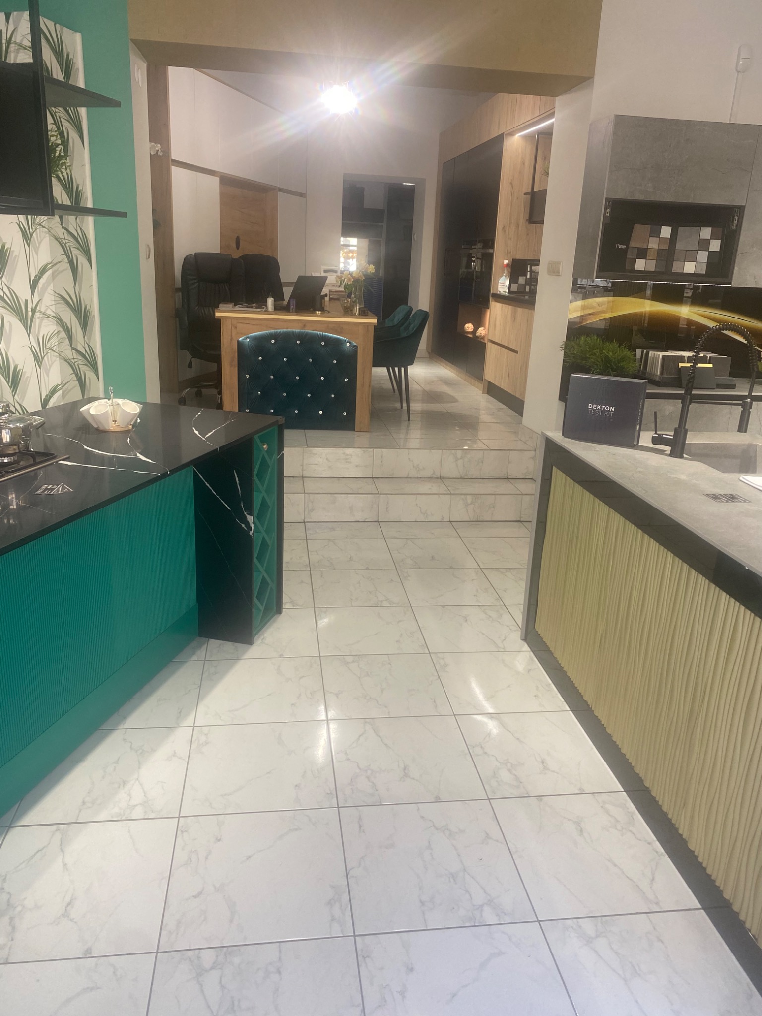 Nowoczesne wnętrze kuchni z wyspą o turkusowym wykończeniu, czarnym blatem i wbudowanym regałem na wino, widok na biuro w tle, białe marmurowe płytki podłogowe.