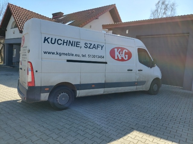Biały samochód dostawczy z napisem 'Kuchnie, Szafy' oraz logo firmy K&G, zaparkowany na brukowanej nawierzchni przed domem z garażem.