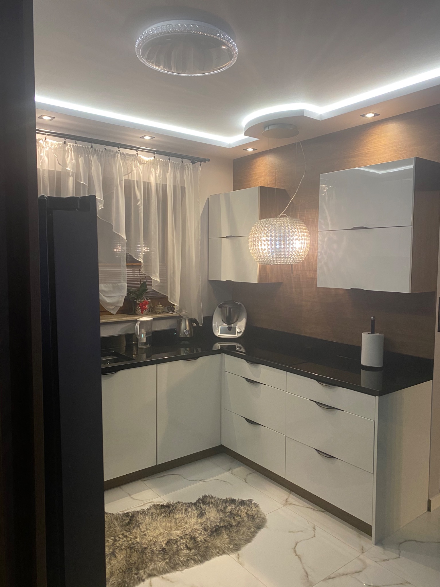 Nowoczesna kuchnia z białymi szafkami, czarnym blatem i drewnianą ścianą, oświetlona ozdobną lampą wiszącą i dywanem na marmurowej podłodze.