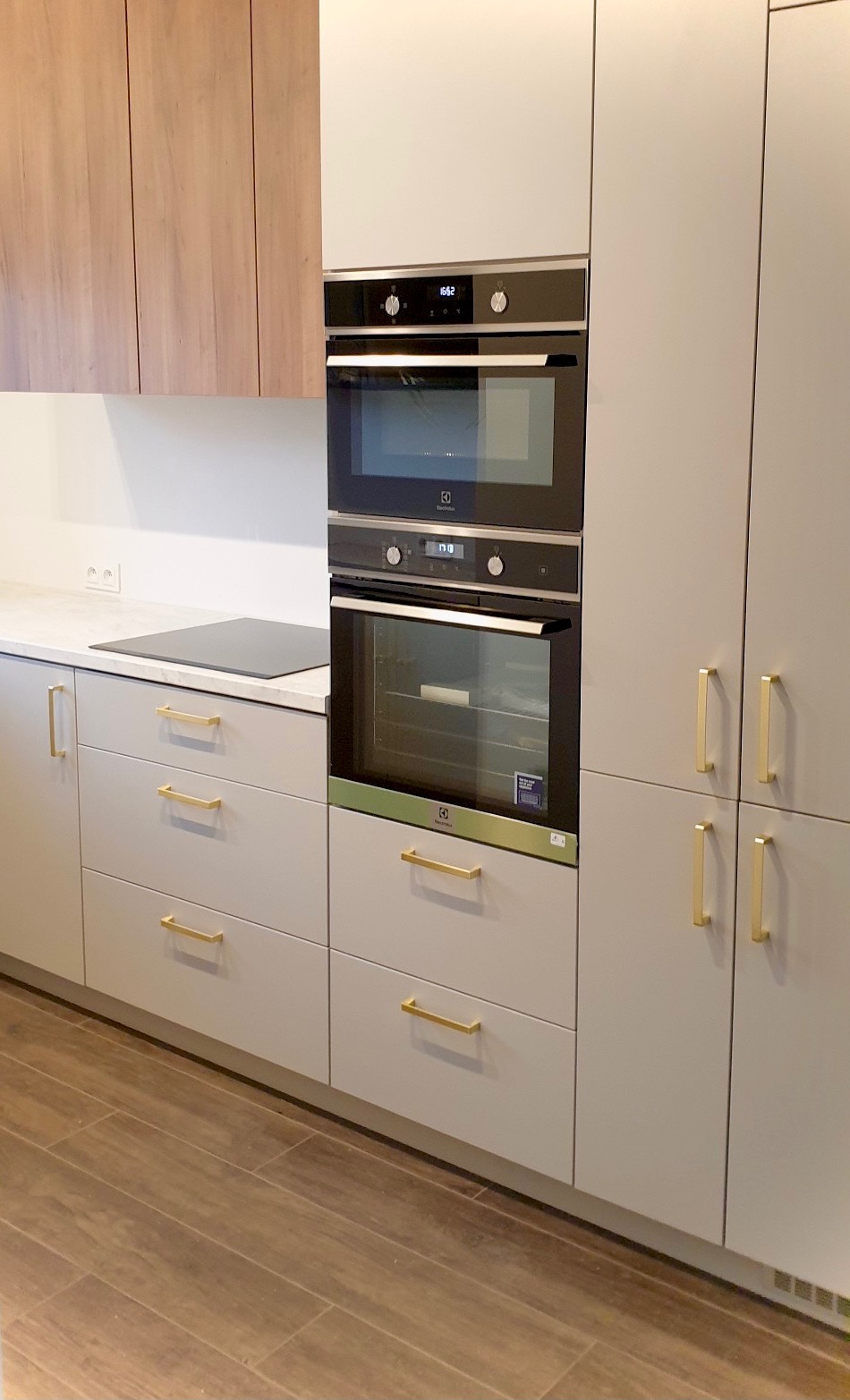 Jasnoszara zabudowa kuchenna z drewnianymi akcentami, piekarnikiem i mikrofalówką Electrolux, oraz złotymi uchwytami, na tle drewnopodobnej podłogi.