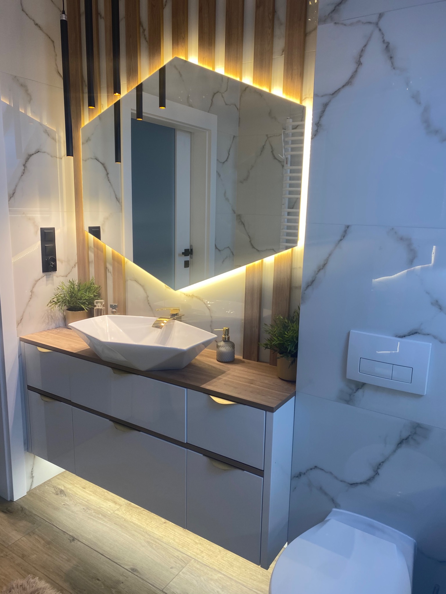 Nowoczesna łazienka z umywalką w kształcie diamentu, podświetlanym lustrem o heksagonalnym kształcie i drewnianymi akcentami na ścianie, marmurowe płytki.