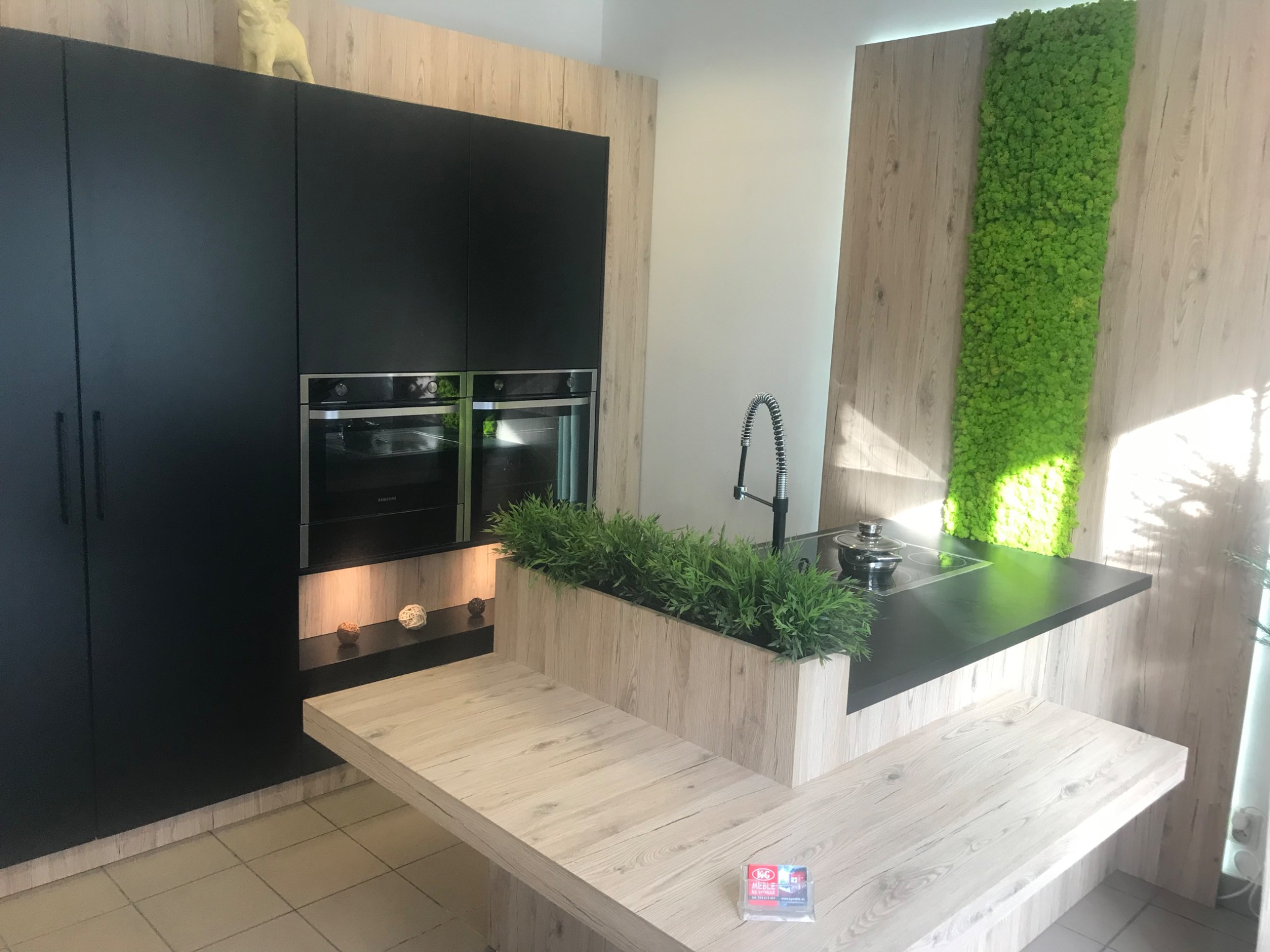 Nowoczesna aranżacja kuchni w Krakowie z czarnymi szafkami, drewnopodobnymi frontami, wyspą z roślinami i panelem z mchu na ścianie.