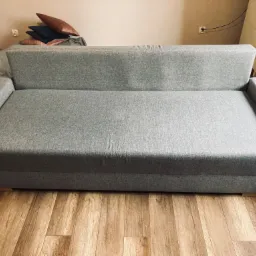 Szara, rozłożona kanapa z poduszkami w tle, stojąca na panelach podłogowych.