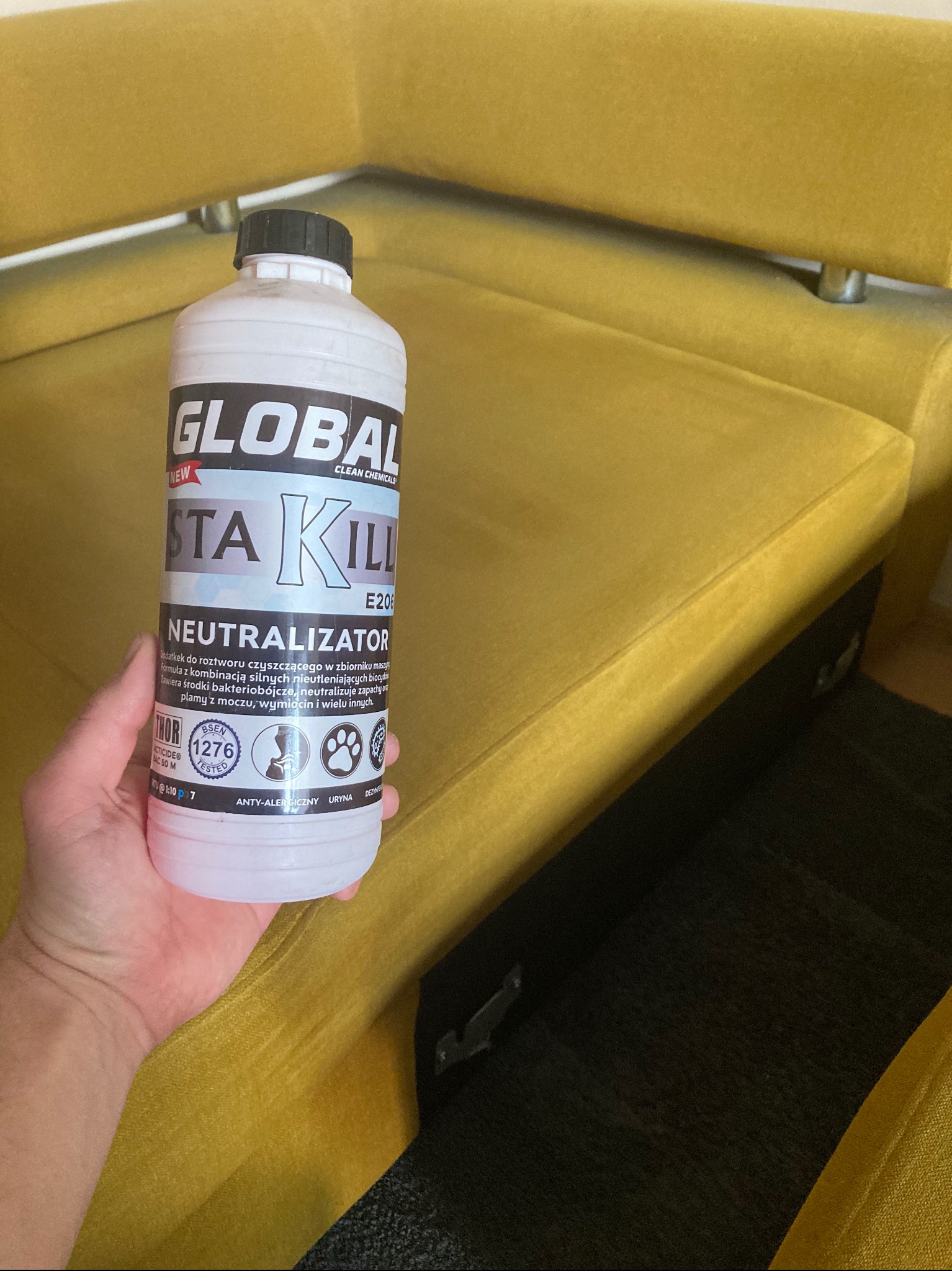 Butelka środka neutralizującego zapachy i plamy, marki Global Sta Kill E206, trzymana w dłoni na tle żółtej, welurowej kanapy.