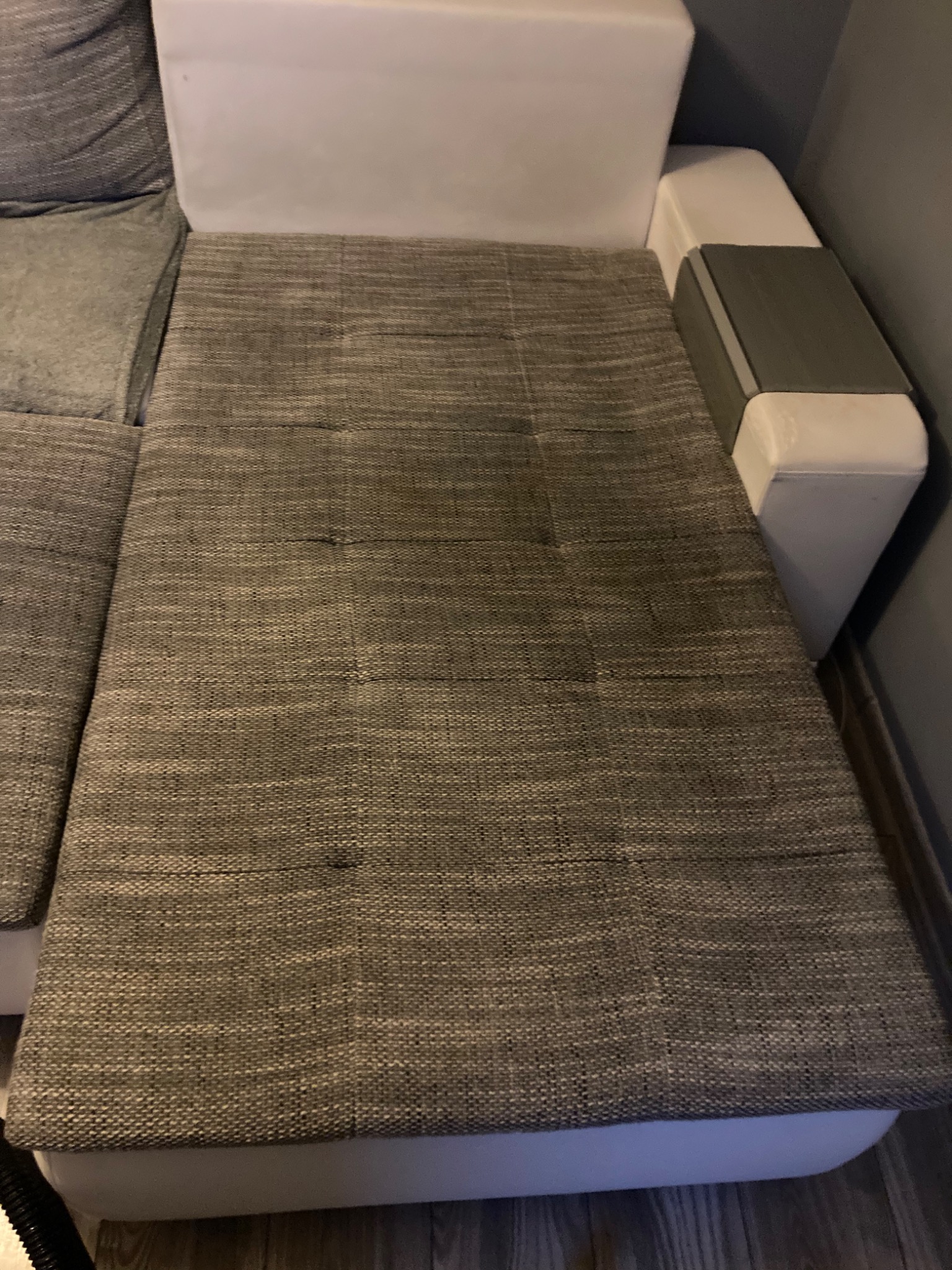 Fragment sofy z szarą teksturowaną tapicerką, widoczne plamy na siedzisku, jasne boki i oparcie.