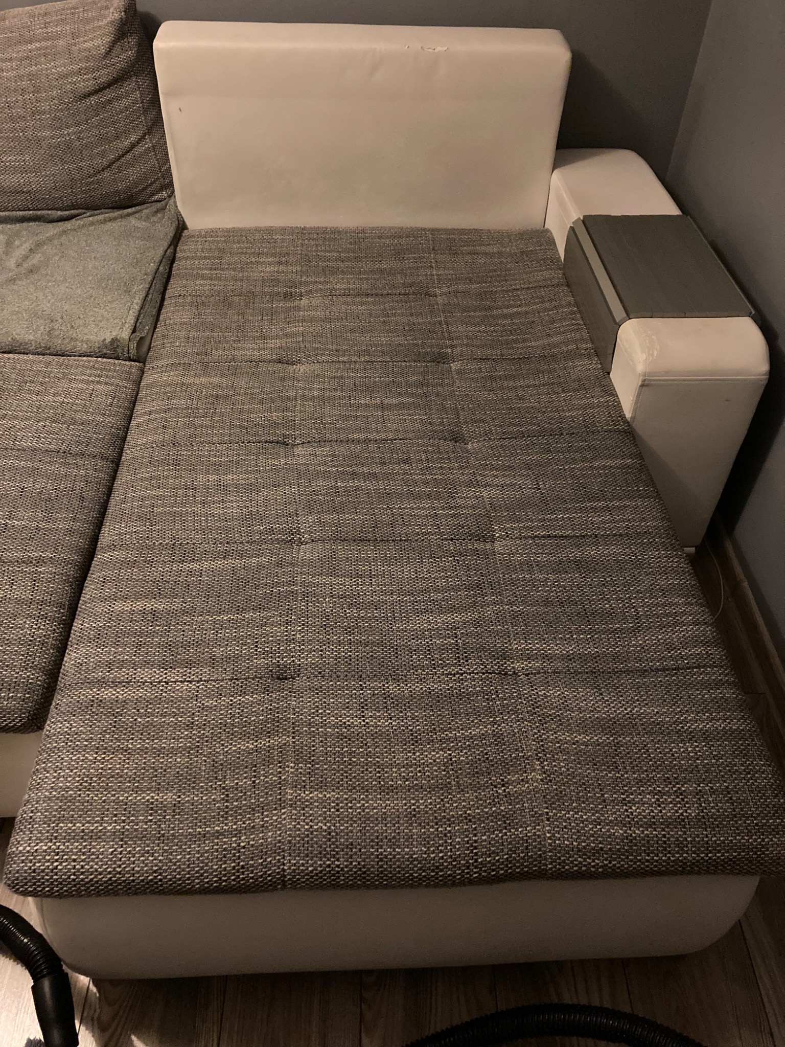 Fragment szarej kanapy narożnej z widocznymi plamami na tapicerce, białe elementy skórzane, czarna rura od odkurzacza w dolnym rogu.