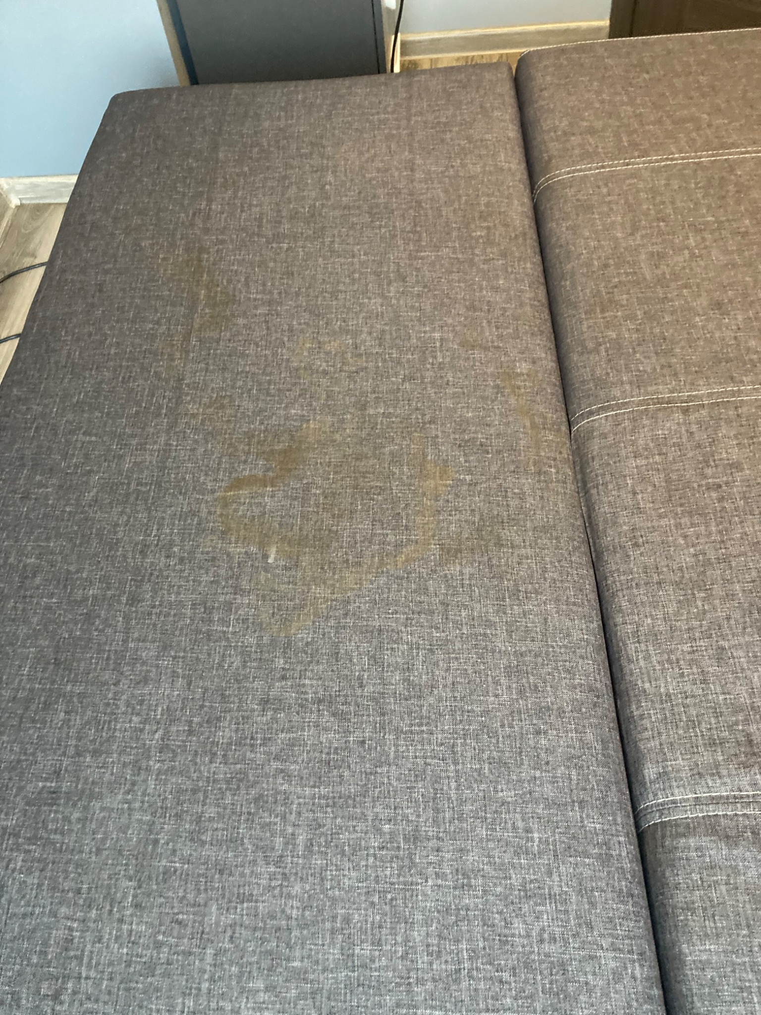 Szara kanapa z wyraźną, okrągłą plamą na siedzisku, widoczne szwy łączące elementy tapicerki.