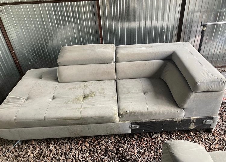 Szara, modułowa kanapa z widocznymi plamami na siedzisku, ustawiona na kamienistym podłożu, sfotografowana w pomieszczeniu z blachy falistej.