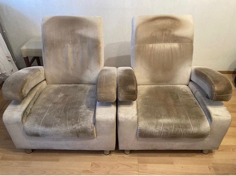 Dwa beżowe fotele tapicerowane z widocznymi, wyraźnymi plamami i zabrudzeniami na siedziskach, oparciach i podłokietnikach, ustawione obok siebie na drewnianej podłodze.