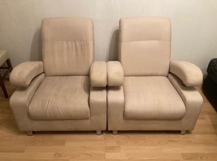 Dwa beżowe, połączone fotele tapicerowane z wysokimi oparciami na drewnianej podłodze, widoczne ślady użytkowania na siedziskach.