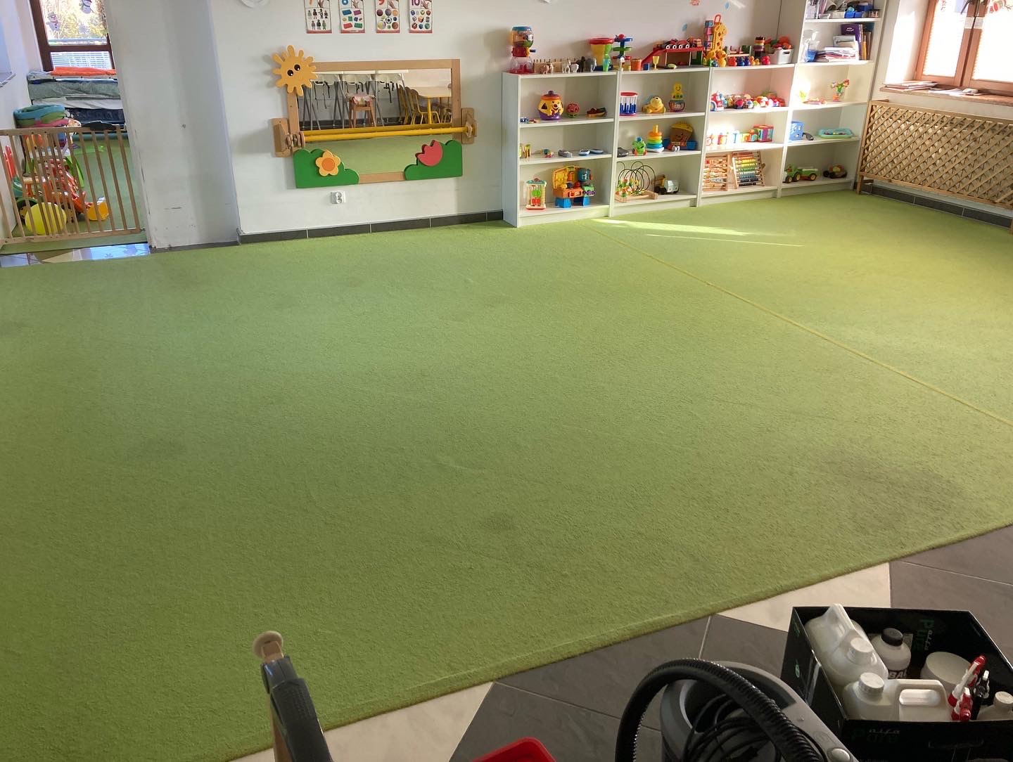 Jasnozielony dywan w pokoju dziecięcym z białymi regałami pełnymi zabawek, lustro z motywami słonecznymi i kwiatowymi na ścianie, w rogu widoczny odkurzacz i pojemnik z środkami czyszczącymi.