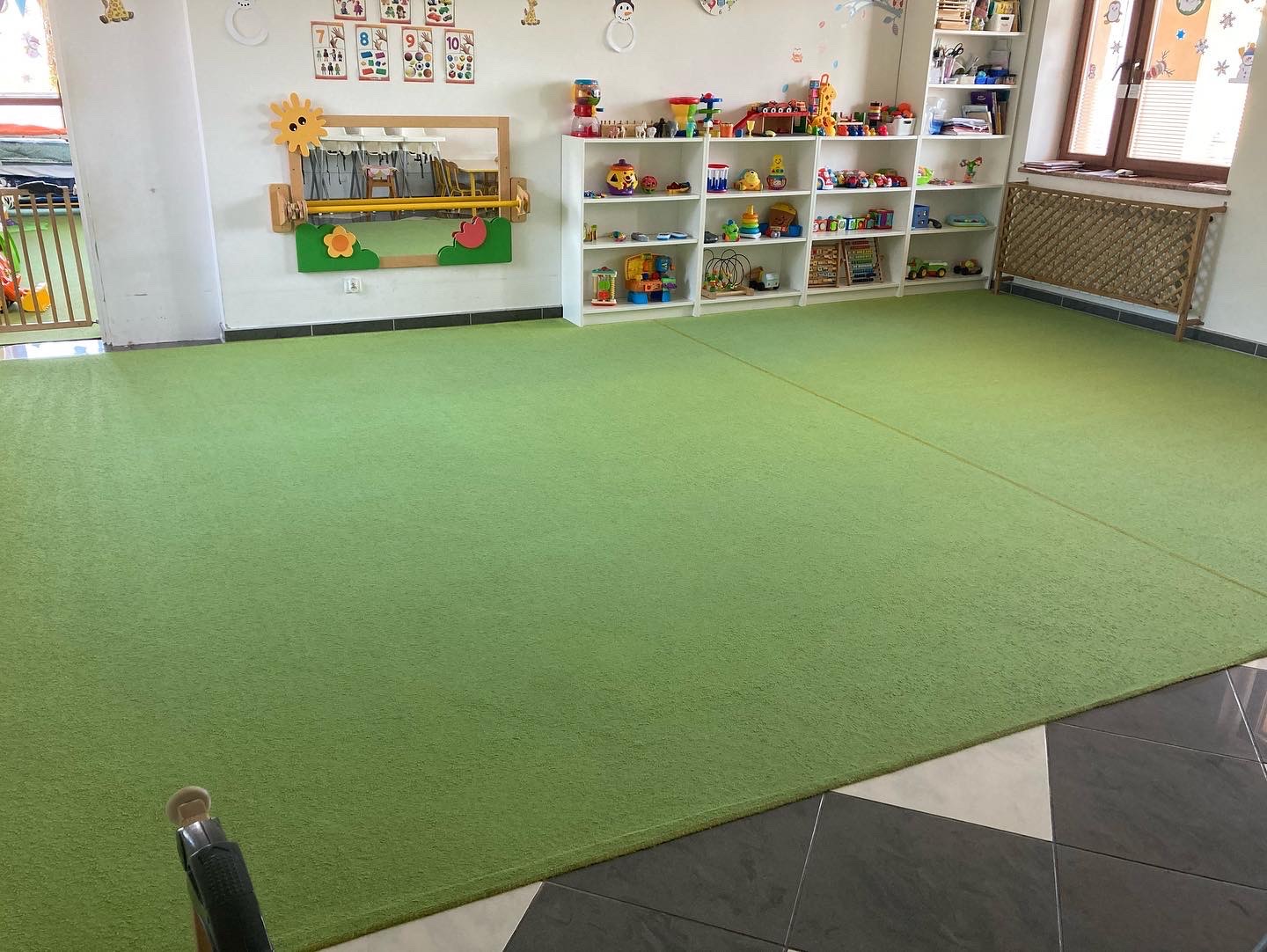 Jasnozielony dywan w pokoju zabaw dla dzieci, z półkami pełnymi zabawek i dekoracjami ściennymi, widoczny fragment czarnego urządzenia do czyszczenia dywanów w dolnym rogu.