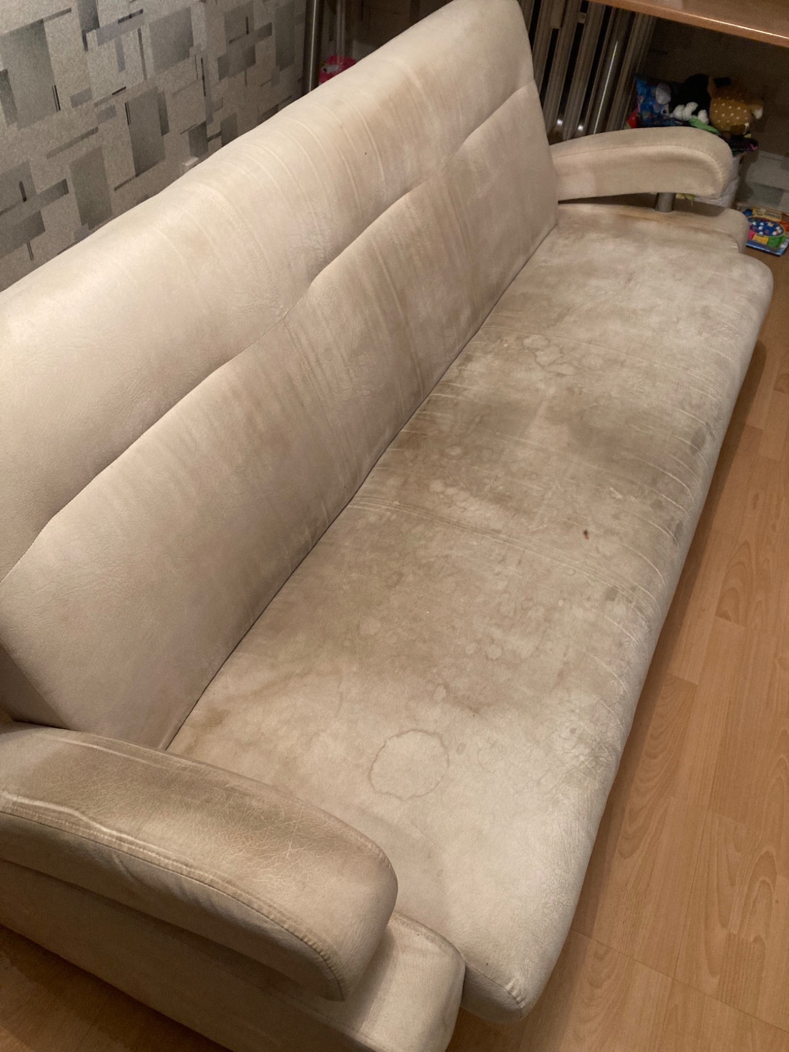 Beżowa kanapa z widocznymi plamami na siedzisku i podłokietnikach, ustawiona na panelach podłogowych.