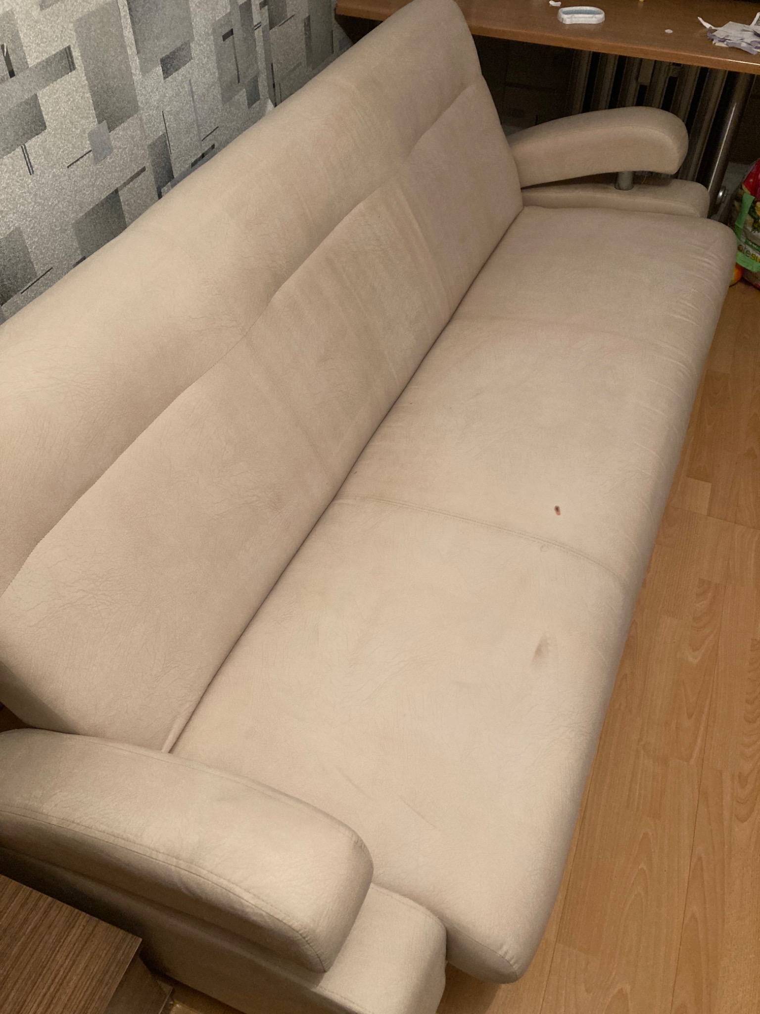 Jasnobeżowa kanapa z widocznymi plamami na siedzisku, ustawiona na tle drewnianej podłogi i ściany z geometrycznym wzorem.