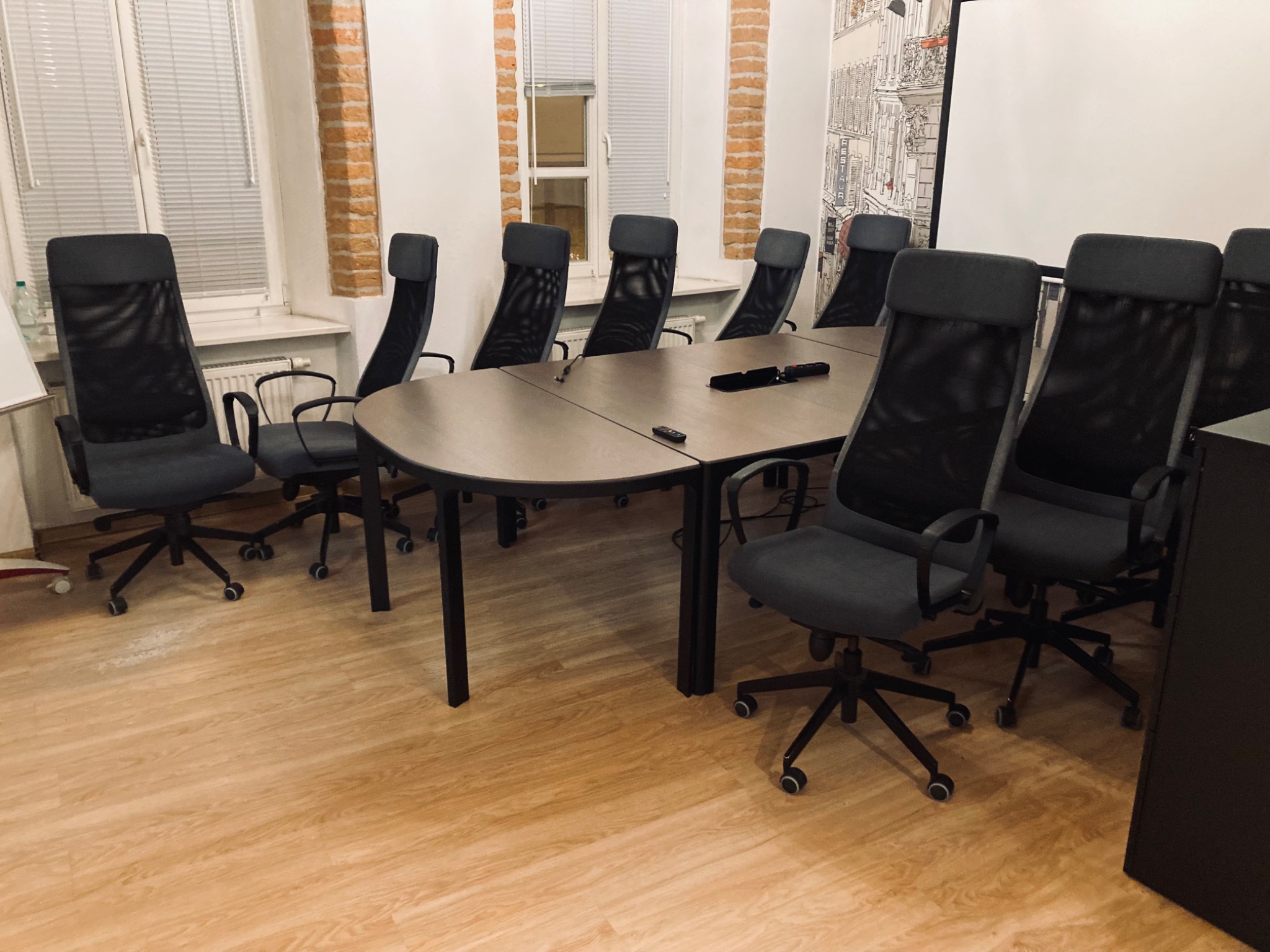 Sala konferencyjna z owalnym stołem i ergonomicznymi krzesłami z siatkowym oparciem, widok na okna z żaluzjami i ścianę z motywem paryskim.