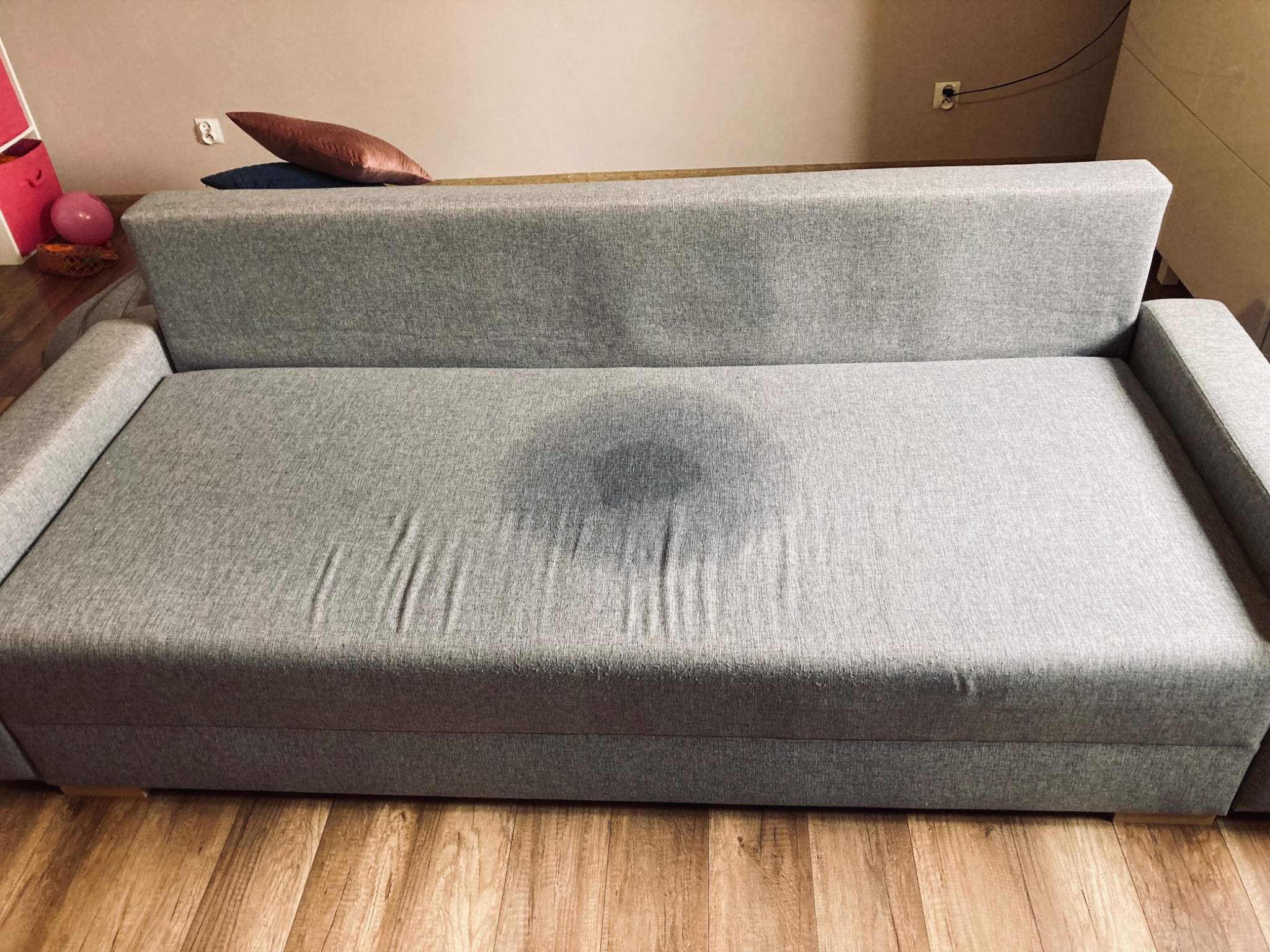 Szara kanapa z widoczną, ciemniejszą plamą na siedzisku, sugerującą świeżo zakończone pranie tapicerki w trakcie schnięcia na tle drewnianej podłogi.