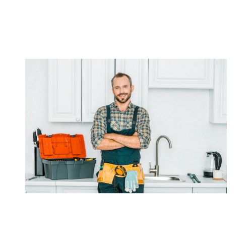Uśmiechnięty mężczyzna w kombinezonie i pasem z narzędziami, stojący na tle zlewu z baterią i szafek kuchennych, obok skrzynki z narzędziami.