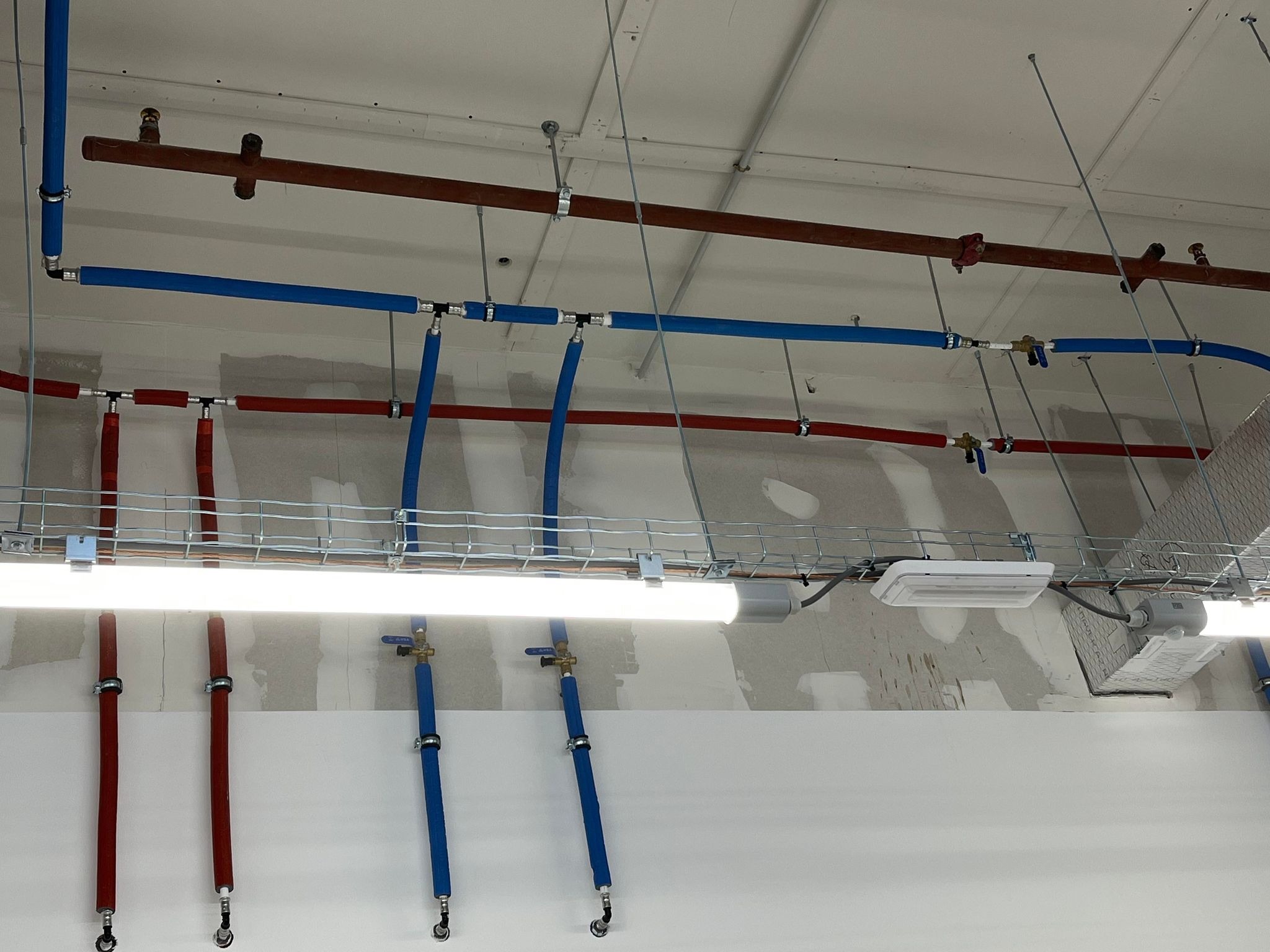 Instalacja wodna z rur PEX w kolorze niebieskim i czerwonym, zamocowana pod sufitem, widoczne zawory kulowe i metalowe obejmy, oświetlenie na metalowej siatce kablowej.