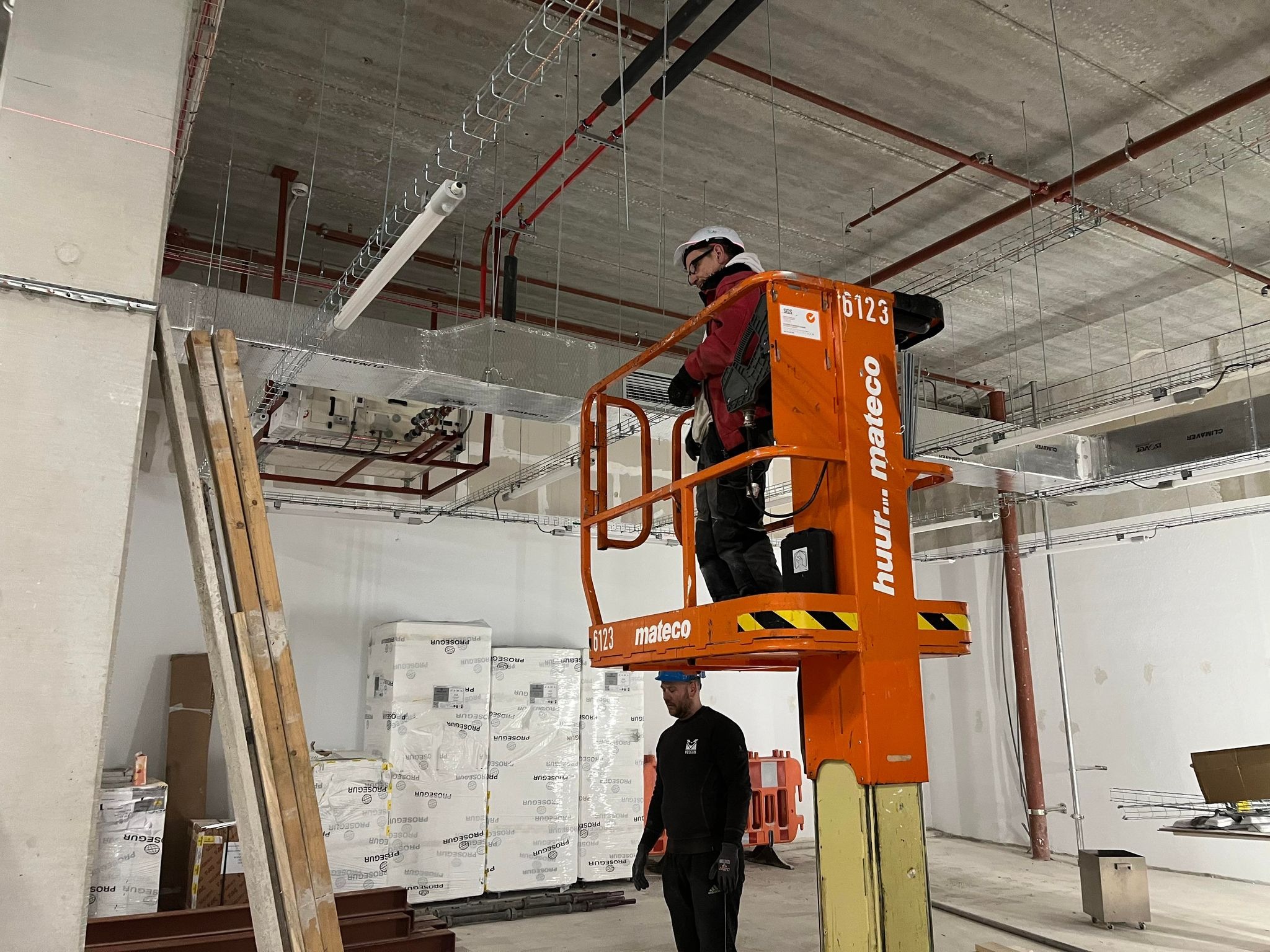 Pracownik w czerwonej kurtce na podnośniku montuje instalacje pod sufitem w Rotterdamie, widoczne rury i korytka kablowe, drugi pracownik obserwuje z dołu.