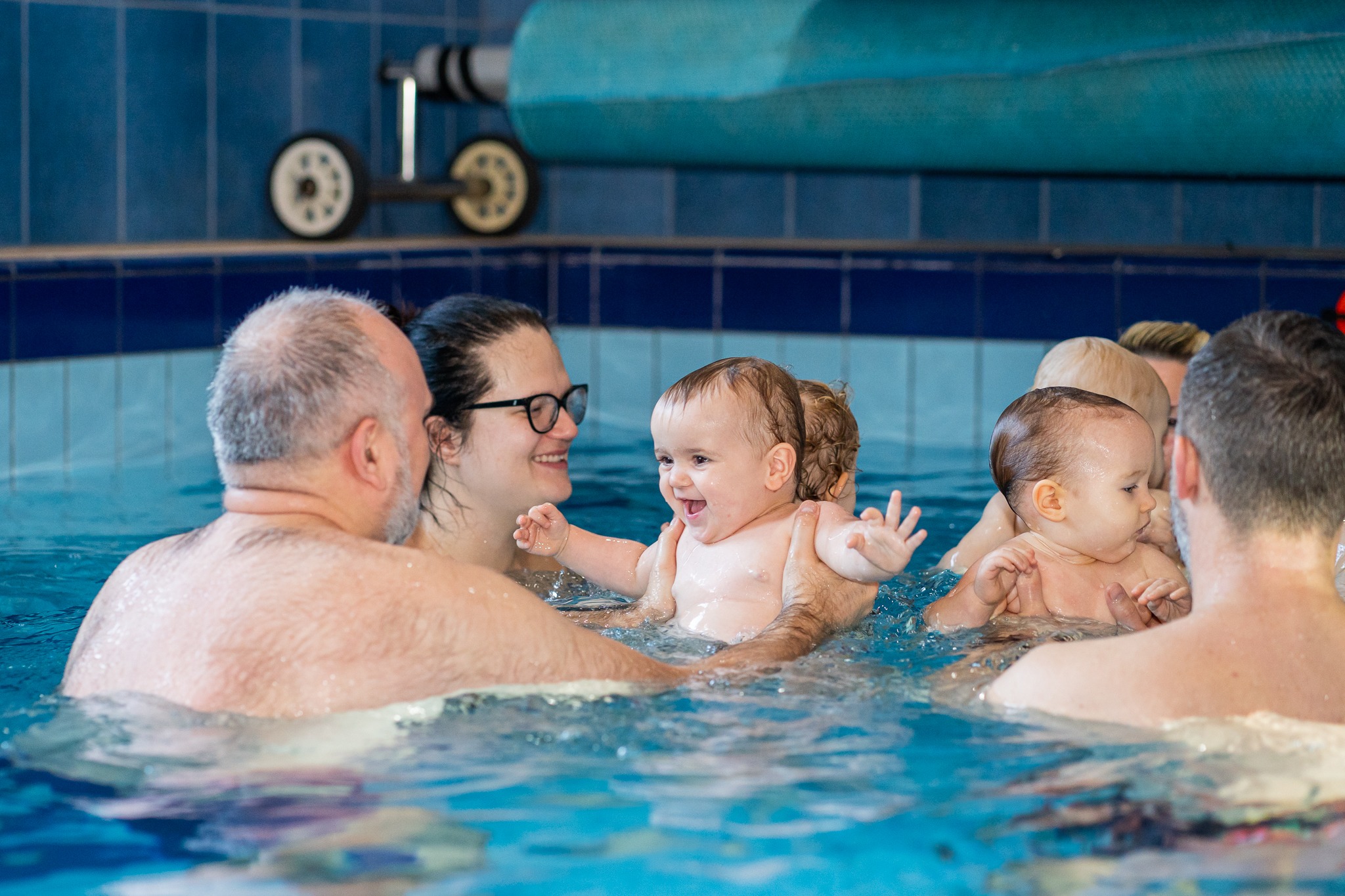 Uśmiechnięte niemowlę trzymane w basenie przez dorosłego, z widoczną kobietą w okularach i innymi dziećmi w tle, na tle niebieskich płytek i zwiniętej pokrywy basenu.
