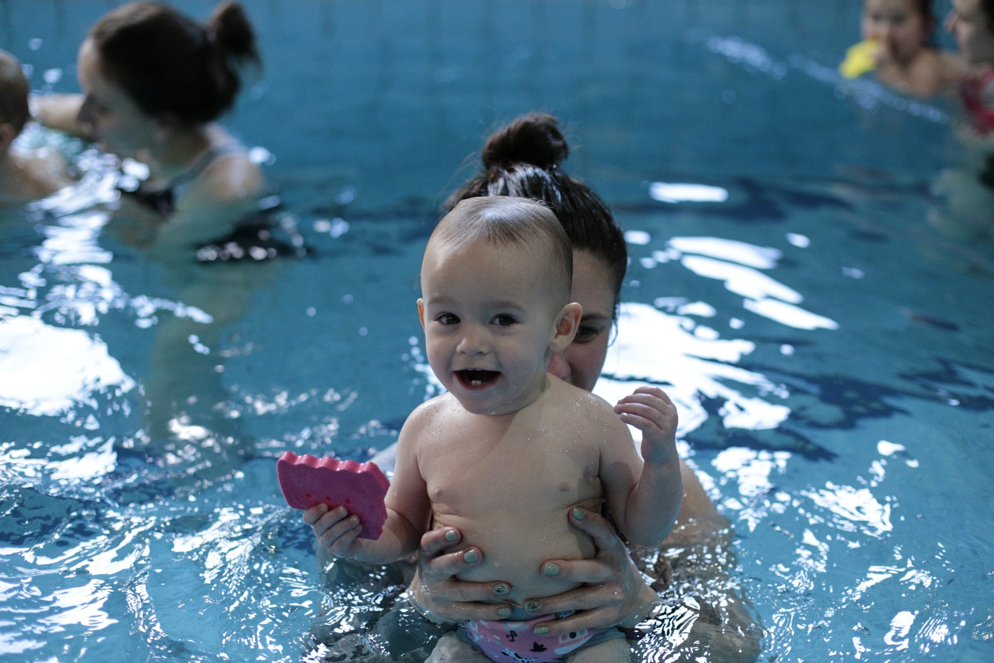 Uśmiechnięte niemowlę trzymane przez dorosłego w basenie, woda odbija światło, dziecko trzyma różową piankową zabawkę, w tle widać inne osoby w wodzie.