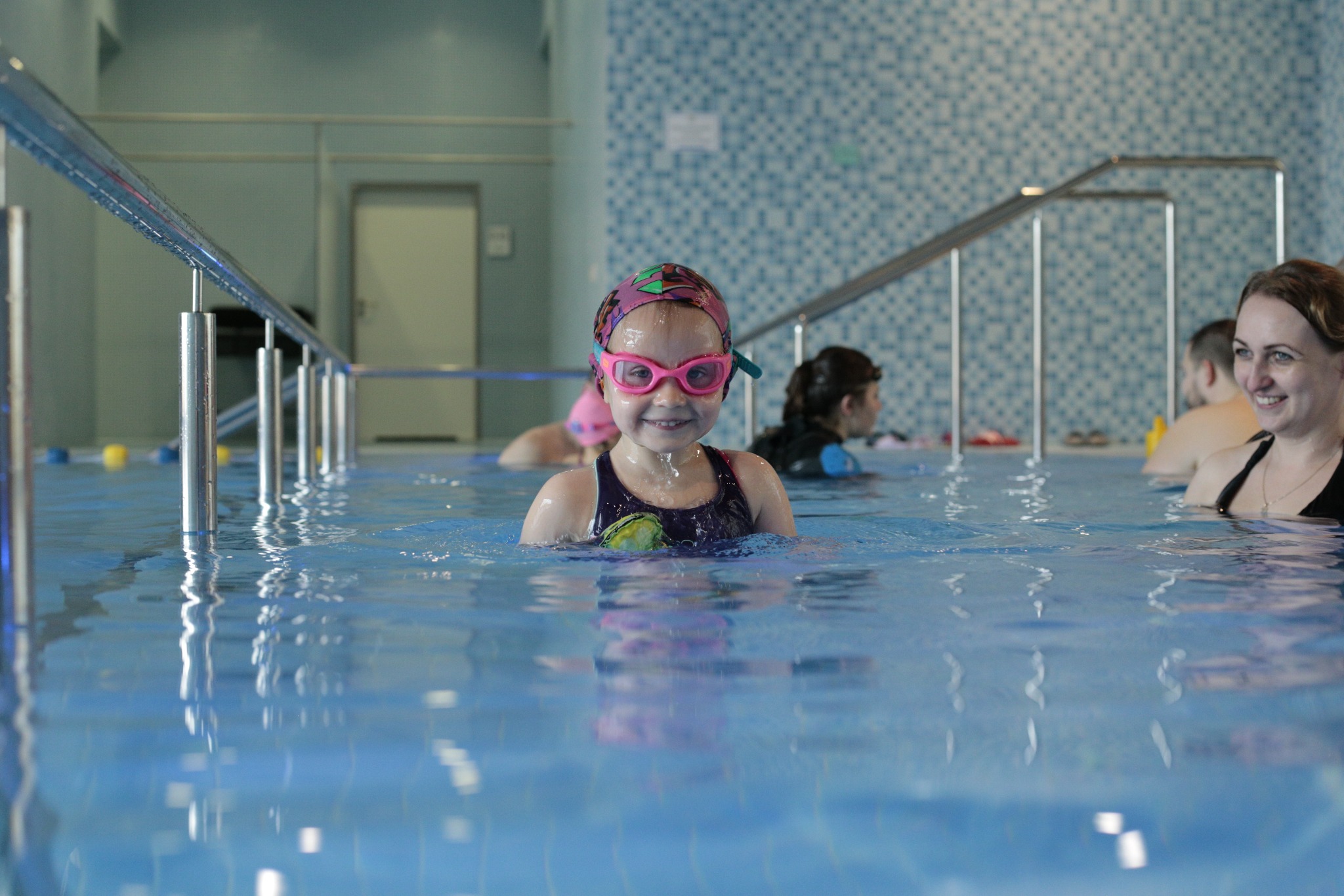 Uśmiechnięta dziewczynka w różowych okularach i czepku pływackim, zanurzona po ramiona w basenie, z instruktorką i innymi uczestnikami w tle.