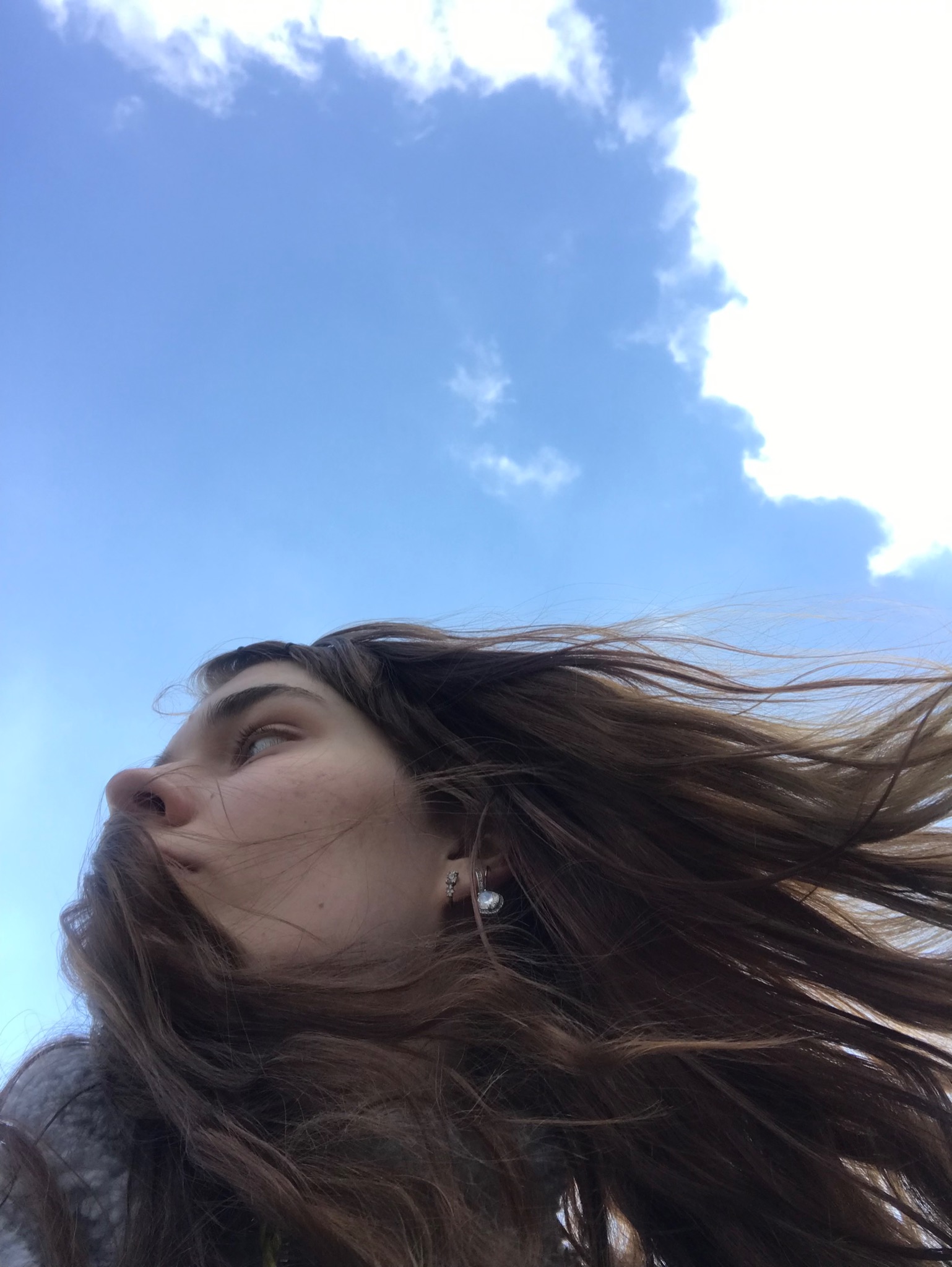 Kobieta z rozwianymi, brązowymi włosami spogląda w górę na błękitne niebo z chmurami, widoczne srebrne kolczyki.