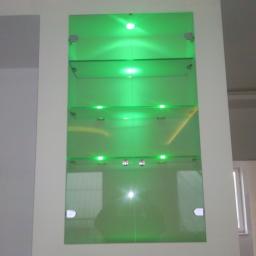 Adrem Adam Banasiak - Szklana witryna z półkami podświetlonymi zielonymi diodami LED, zamontowana we wnęce ściany.