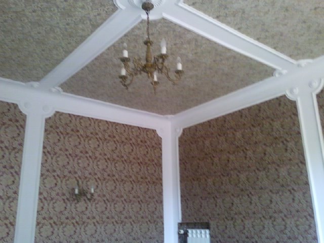 Pokój z tapetowanymi ścianami w brązowe wzory, białe listwy dekoracyjne na suficie i ścianach, oraz ozdobny żyrandol.