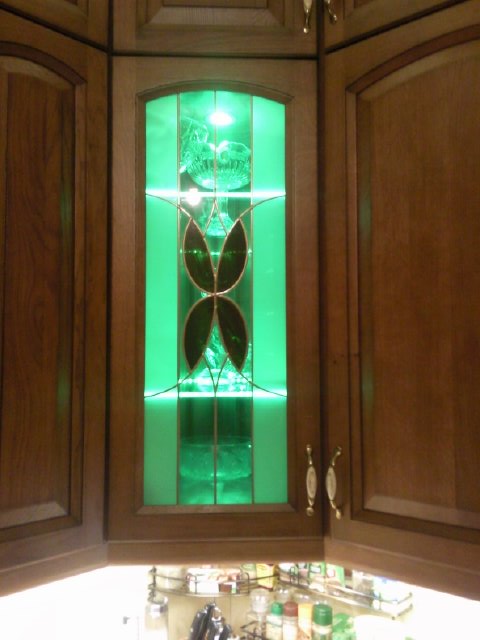 Drewniana szafka narożna z witrażem oświetlonym od wewnątrz zielonym światłem LED, prezentująca szklane naczynia.