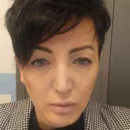 Portret kobiety z krótkimi, ciemnymi włosami w marynarce w pepitkę, patrzącej prosto w obiektyw.