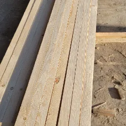 Stos jasnej, świeżo obrobionej tarcicy na placu budowy, widoczne słoje drewna i drobne wióry.