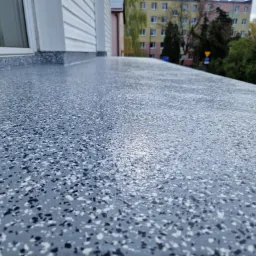 Szara, błyszcząca powierzchnia balkonu wykończona żywicą z zatopionymi płatkami, widok z niskiej perspektywy, w tle budynki mieszkalne.