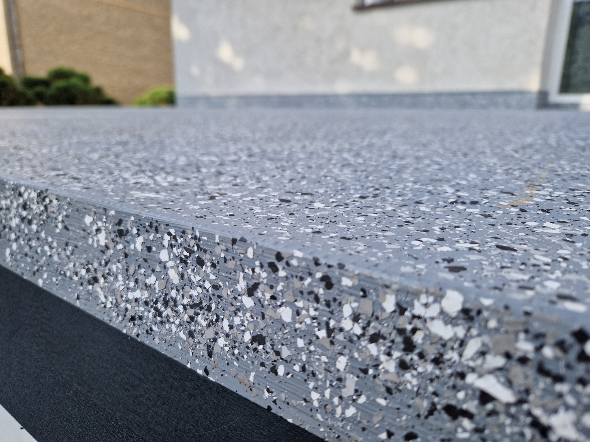 Szara, gładka powierzchnia z widoczną strukturą żywicy epoksydowej z zatopionymi płatkami w odcieniach szarości, bieli i czerni, tworząca wykończenie podłogi na zewnątrz budynku.