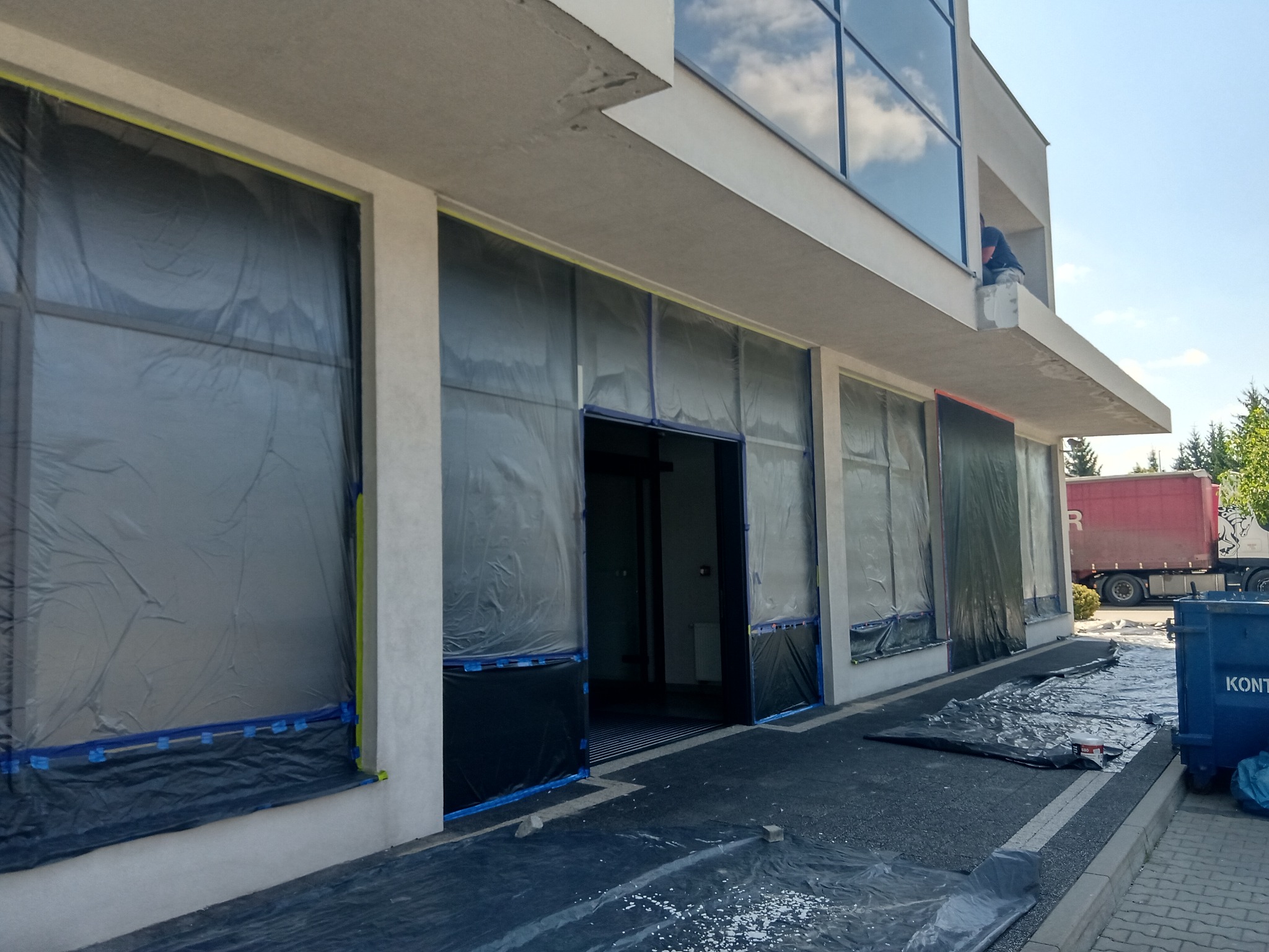 Przygotowanie budynku do malowania: okna i drzwi zabezpieczone folią malarską, ślady farby na chodniku, widoczny pracownik na balkonie.